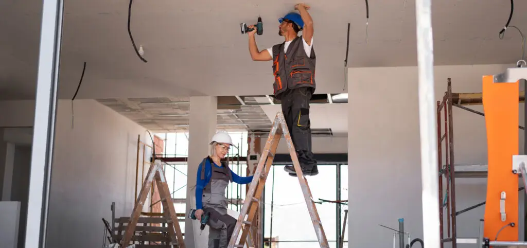 Two construction workers on ladders install cables on a ceiling inside a building under renovation, with tools and equipment visible around them.