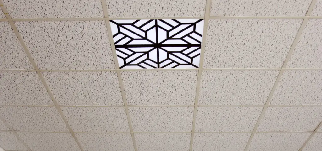 A suspended ceiling with white acoustic tiles features one central tile with a geometric patterned vent.