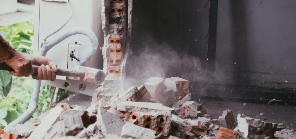 A person uses a sledgehammer to demolish a brick wall, creating dust and debris with broken bricks scattered on the ground.