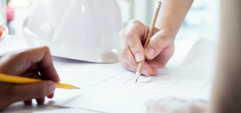 Two people hold pencils and review architectural blueprints on a desk, with a white hard hat in the background.